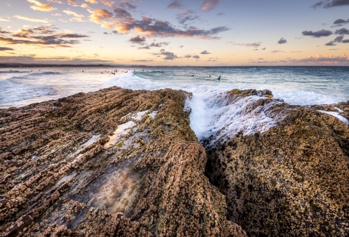 Waves crashing over rocks with surfers in the background at sunset