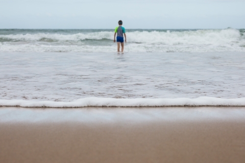 Waves rolling in on sand at beach with young boy walking out to ocean - Australian Stock Image
