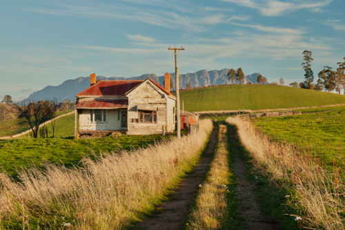 Weathered farmhouse with a rust‑red roof sits quietly beside a narrow dirt road - Australian Stock Image