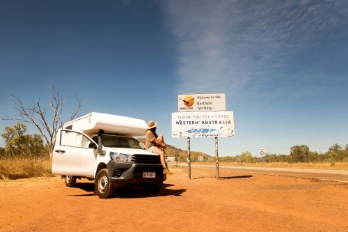 White camper van and a woman sitting on the hood looking at the signage beside her in a rural area