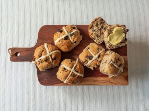 Whole and cut Hot Cross Buns with butter on wooden serving board on white table cloth - Australian Stock Image