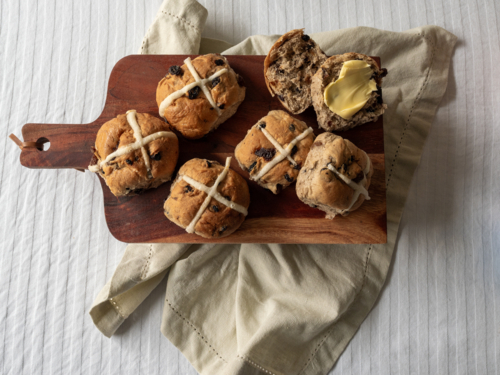 Whole and cut hot cross buns with butter on wooden serving tray with white table cloth, linen napkin - Australian Stock Image