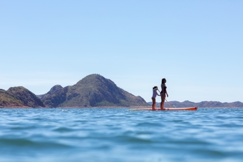 woman and child standing on paddle board in blue water with hills behind - Australian Stock Image
