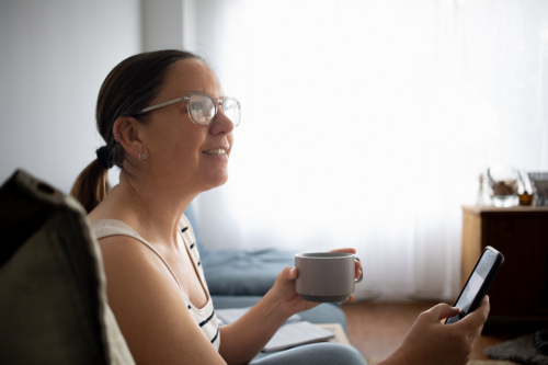Woman at home looking at someone while using smart phone and drinking coffee - Australian Stock Image