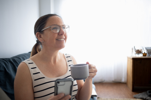 Woman at home on couch smiling at someone while using smart phone and drinking coffee - Australian Stock Image