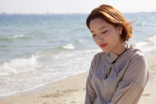 Woman at the beach looking to the ground with the sand and sea behind her - Australian Stock Image