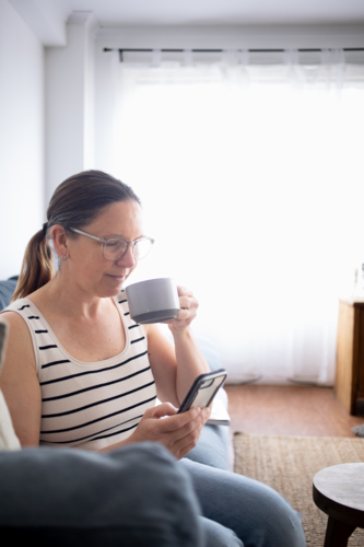 Woman drinking coffee on couch and using smart phone - Australian Stock Image