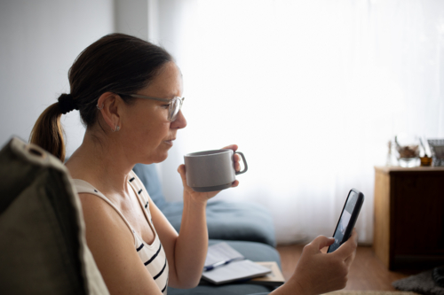 Woman drinking coffee on couch at home while concentrating on smart phone - Australian Stock Image