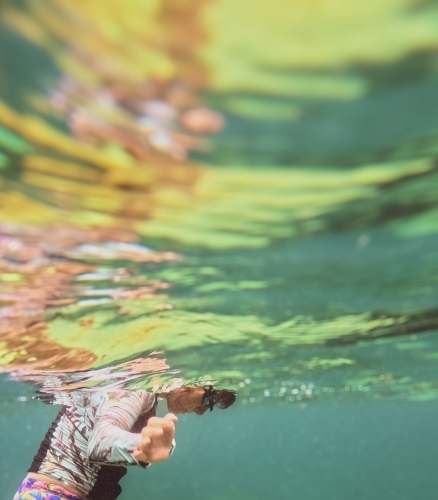 Woman floating and looking underwater with colourful reflection wearing goggles and rash vest - Australian Stock Image