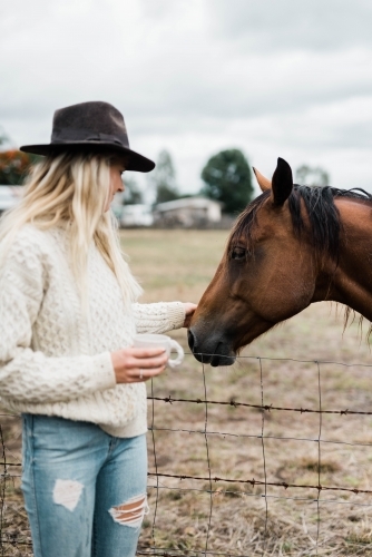 Woman holding a cup of coffee while the pats a horse