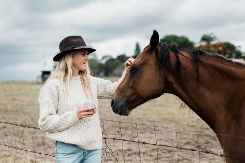 Woman holding a cup of coffee while the pats a horse - Australian Stock Image