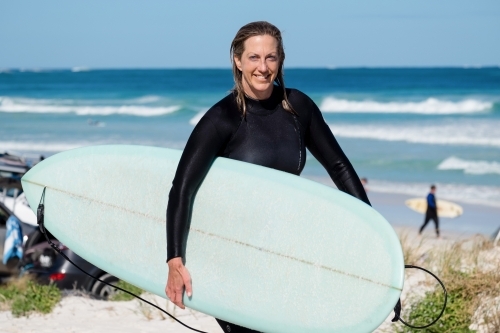 Woman holding longboard under arm in wetsuit with wet hair on coastal dune and ocean in background - Australian Stock Image