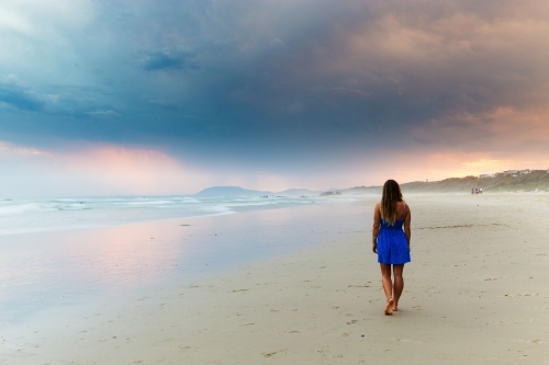 Woman in a blue dress walking on the beach towards a stormy sunset