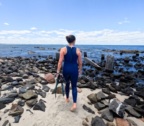Woman in blue wetsuit walking along rocky beach coastline holding snorkelling fins & mask - Australian Stock Image