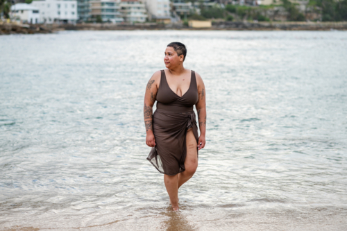 Woman in brown swimsuit walking in shallow waters at the beach - Australian Stock Image
