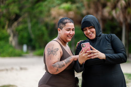 Woman in burkini showing her phone to her friend on the sandy shore at the beach - Australian Stock Image