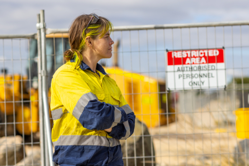 Woman in profile on worksite wearing hi-vis in front of temporary fencing - Australian Stock Image
