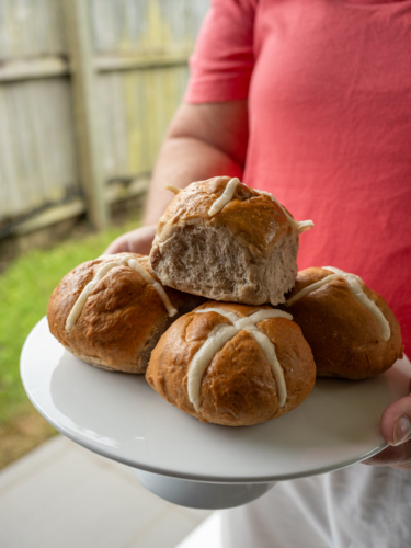 Woman in red shirt holding a cake stand piled with hot cross buns with green grass and wooden fence - Australian Stock Image