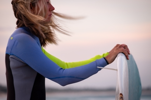 Woman in wetsuit holding surfboard on beach with windswept hair - Australian Stock Image