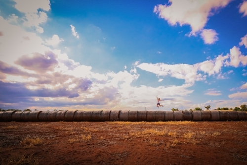 Woman jumping for joy on long row of hay bales on farm - Australian Stock Image