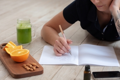 woman lying on floor goal setting in journal with healthy snacks orange almonds green juice phone