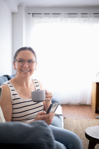 Woman on couch drinking coffee, using smart phone - Australian Stock Image