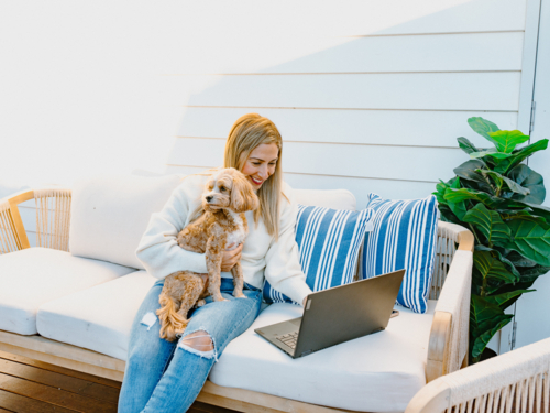 Woman on her laptop while holding a dog on the couch. - Australian Stock Image