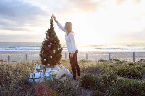 Woman placing decoration on top of Christmas tree on beach at sunrise