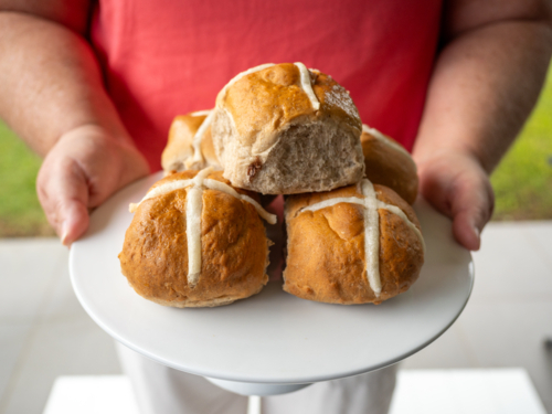 Woman's hands holding white plate piled with hot cross buns in backyard - Australian Stock Image