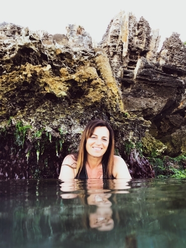 Woman sitting in glassy ocean and cupping water with coastal limestone rocks in background - Australian Stock Image