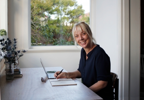 Woman sitting in sun room office working with laptop and notepad - Australian Stock Image