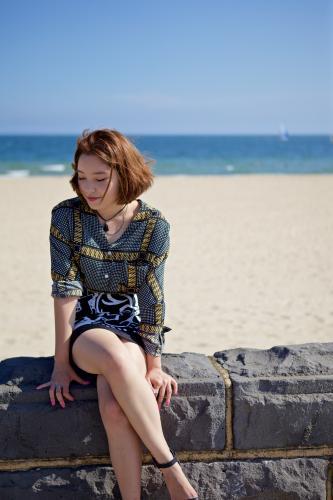 Woman sitting on a bluestone wall with the beach, sand and sea behind her - Australian Stock Image