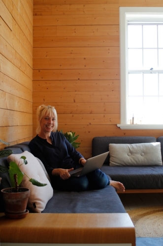 Woman sitting on couch working on laptop with timber wall background - Australian Stock Image