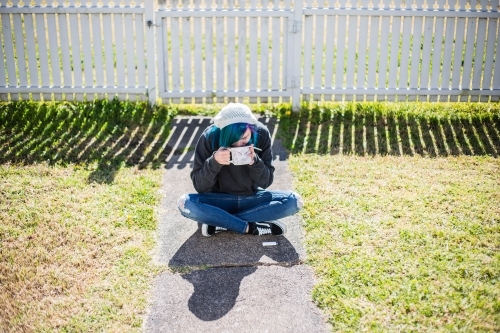 Woman sitting on path in front of white fence drinking coffee and holding smoke