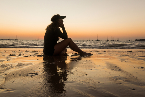 Woman sitting on shoreline listening to music on headphones at sunset - Australian Stock Image