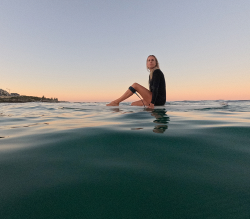 Woman sitting on surfboard floating on glassy water with leg up on board and sunrise sky - Australian Stock Image