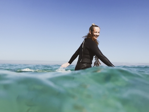 Woman sitting on surfboard paddling arms looking at camera wearing seaweed