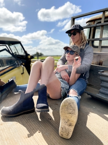 Woman sitting with teenage child cuddled against her on rustic farm truck in work boots - Australian Stock Image