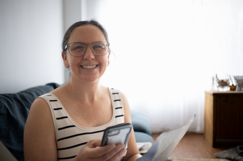 Woman smiling at camera, using smartphone and reading document at home - Australian Stock Image