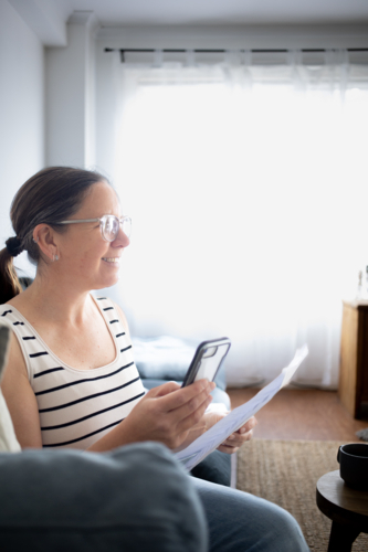 Woman smiling while using smart phone and reading document at home - Australian Stock Image