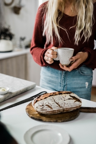 Woman standing behind kitchen bench holding a cup of coffee - Australian Stock Image