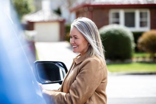 Woman standing beside parked blue car in a parking area smiling - Australian Stock Image