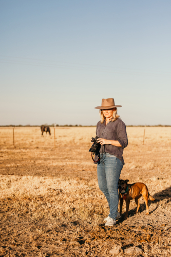 Woman standing in a dusty and dry grassy farm paddock holding a camera - Australian Stock Image