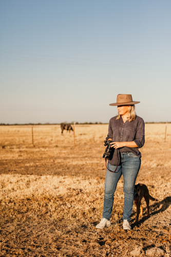 Woman standing in a dusty and dry grassy farm paddock holding a camera - Australian Stock Image