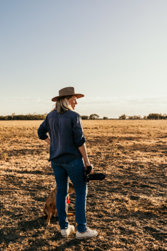 Woman standing in a dusty and dry grassy field holding a camera - Australian Stock Image