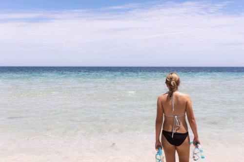 Woman standing in front of clear blue water with snorkelling equipment - Australian Stock Image