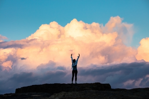 Woman standing with arms up on rock against a dramatic cloud backdrop - Australian Stock Image
