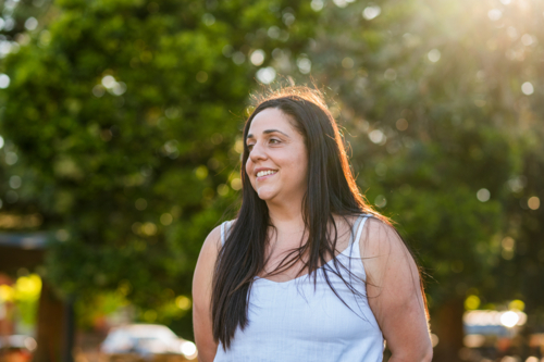 Woman stands smiling in a park surrounded by greenery and bright sunlight during the day - Australian Stock Image
