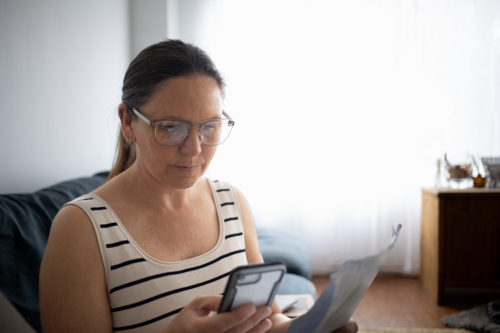 Woman using smartphone and reading document at home - Australian Stock Image