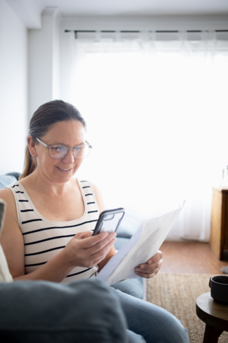 Woman using smartphone and reading document at home - Australian Stock Image